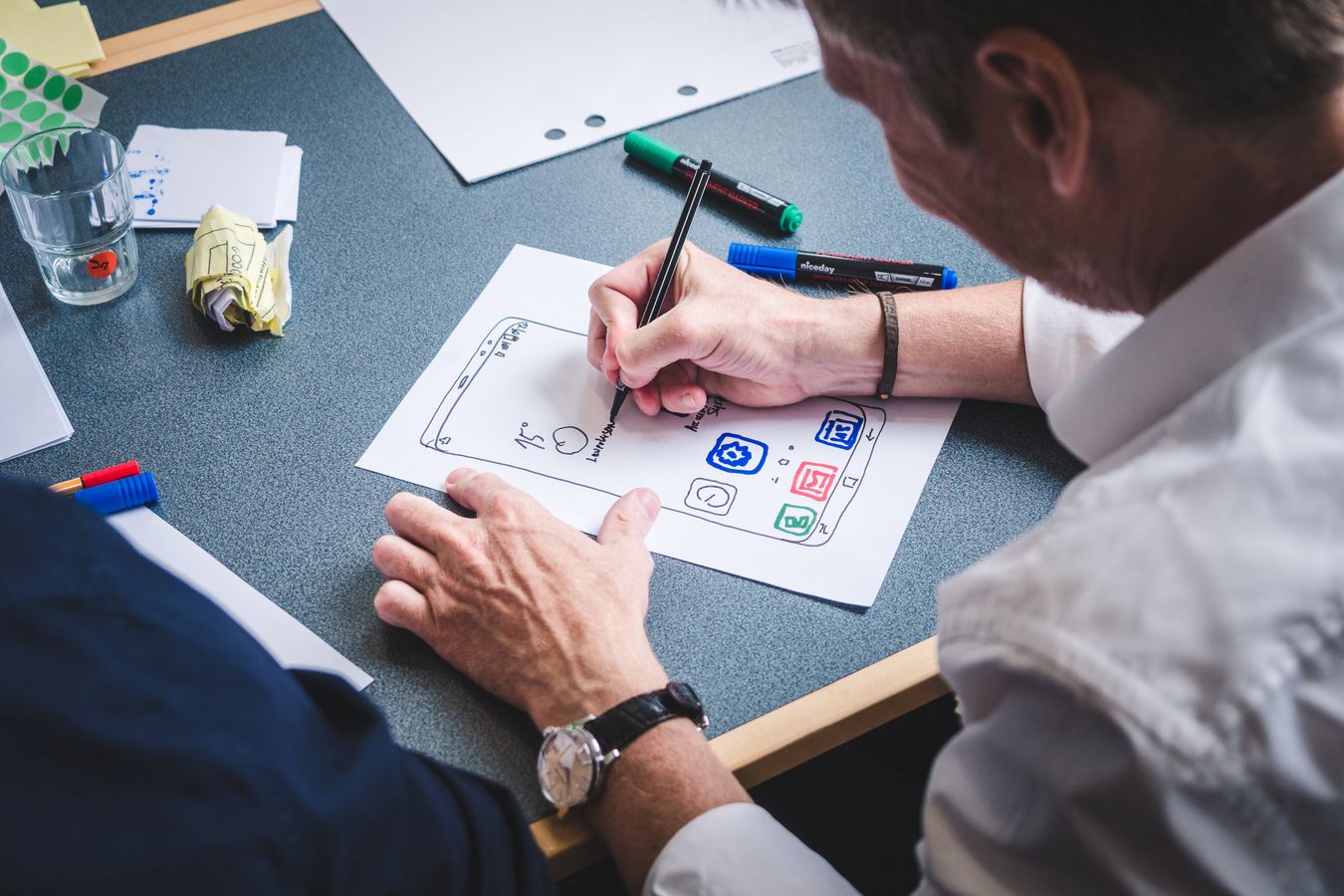 A man sketches a phone app home screen with coloured icons on paper, markers and crumpled drafts on the table