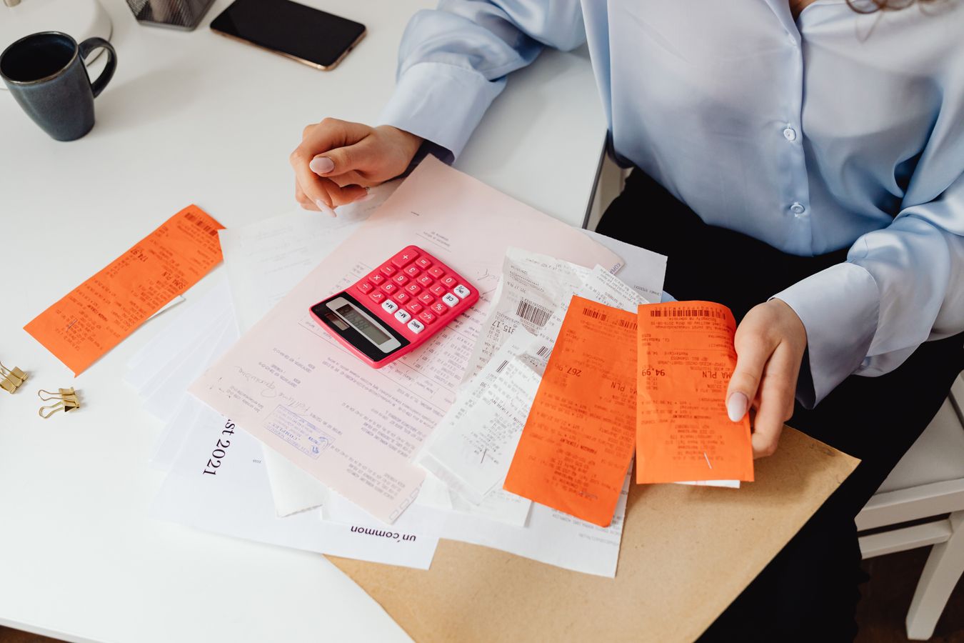 A person at a white desk holds a handful of paper receipts next to a pink calculator and a pile of invoices