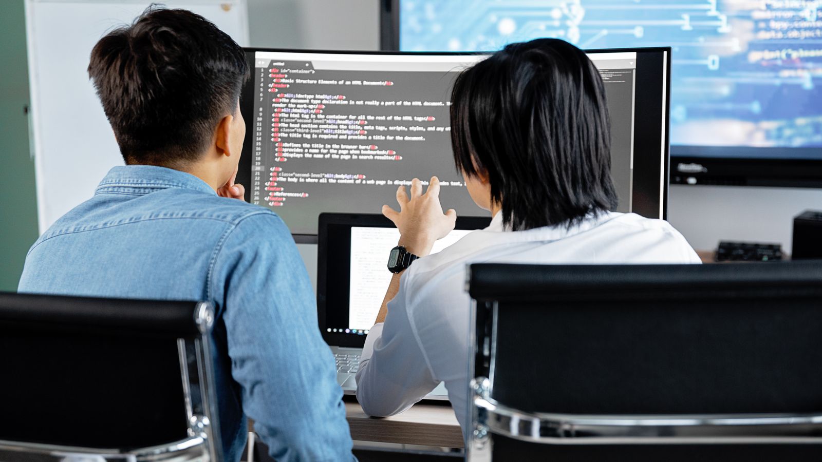 Two developers seen from behind, reading code together on a wide monitor
