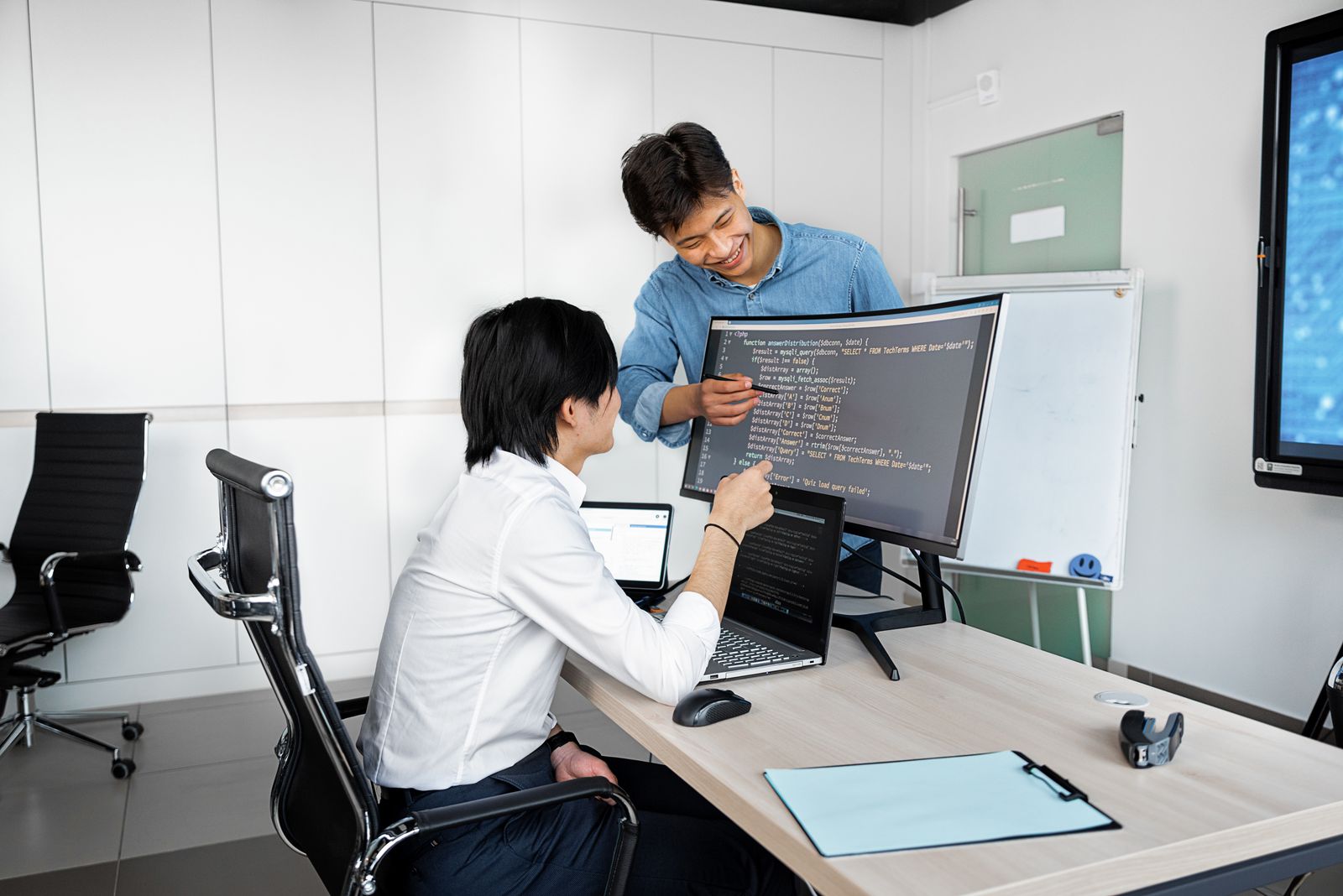 Two developers in an office looking at code on a large curved monitor, one pointing at the screen