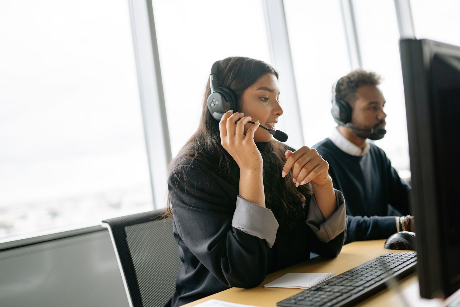 Two customer service agents with headsets taking calls at a desk by a large window