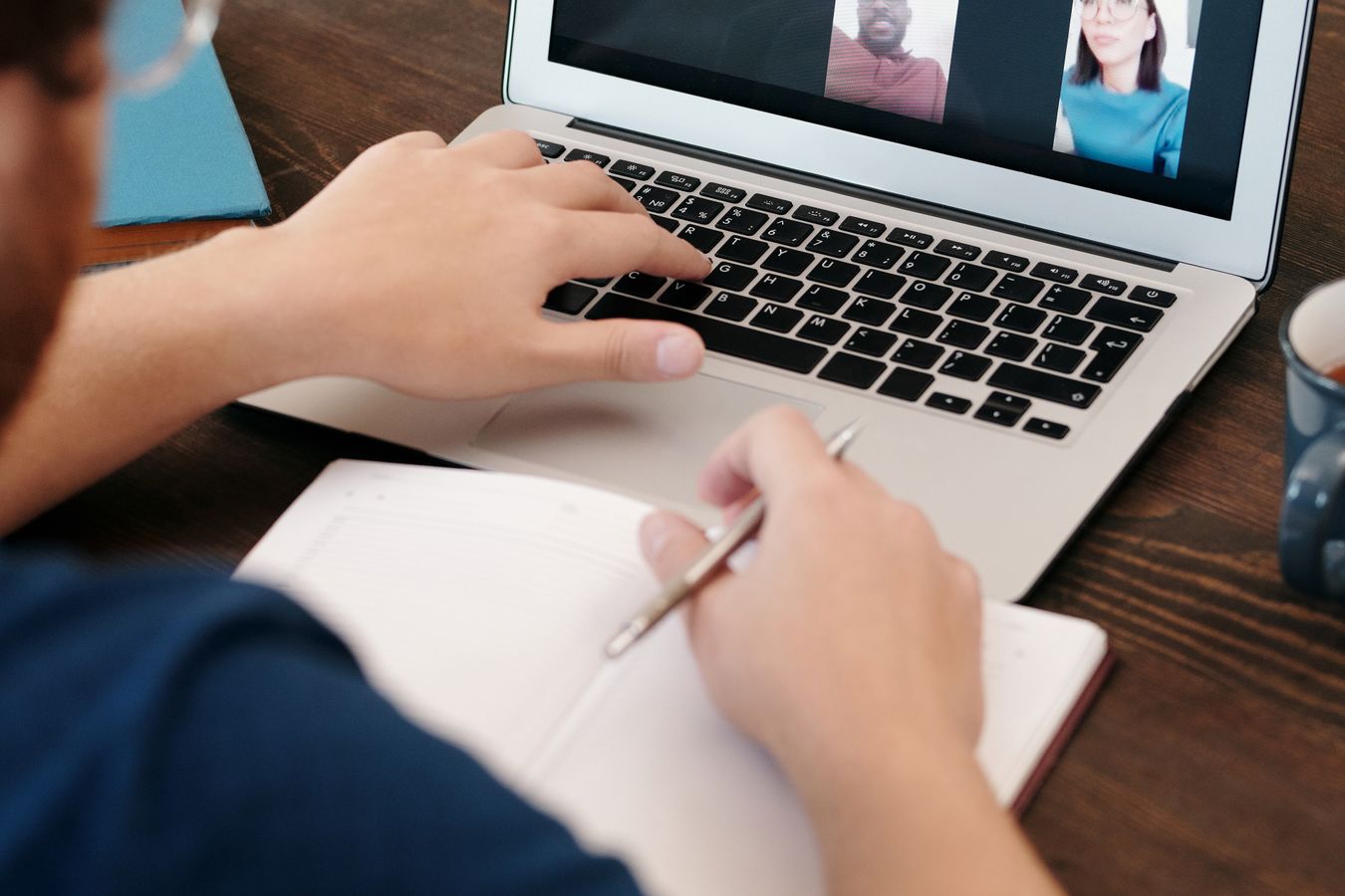 Over the shoulder of a man taking notes in a notebook while two colleagues appear on a video call on his laptop