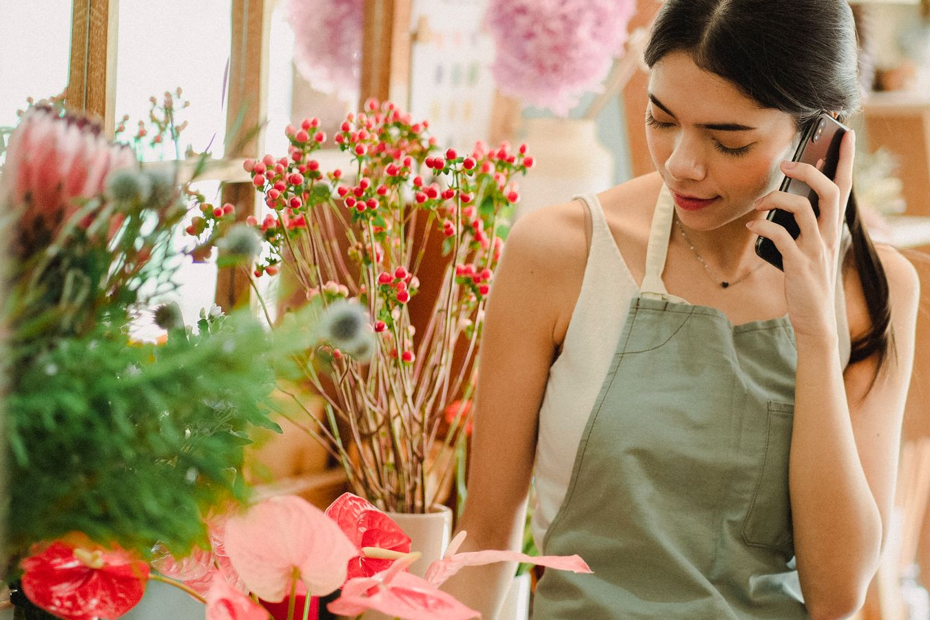 A florist in a green apron takes a call on her mobile phone among buckets of flowers in her shop