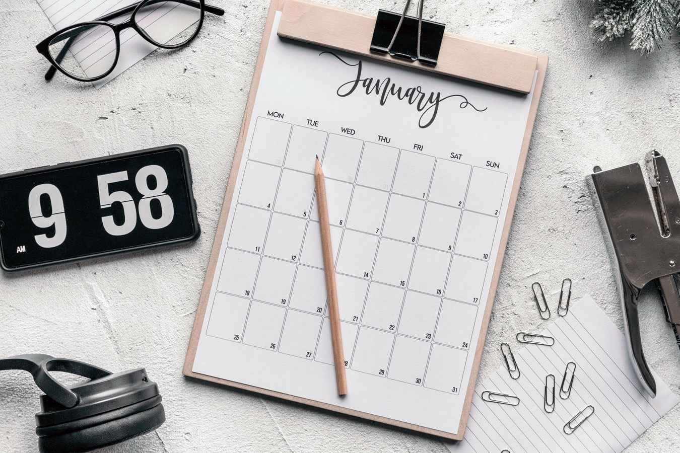 A January calendar page on a wooden clipboard with a pencil, beside a flip clock, glasses and paper clips