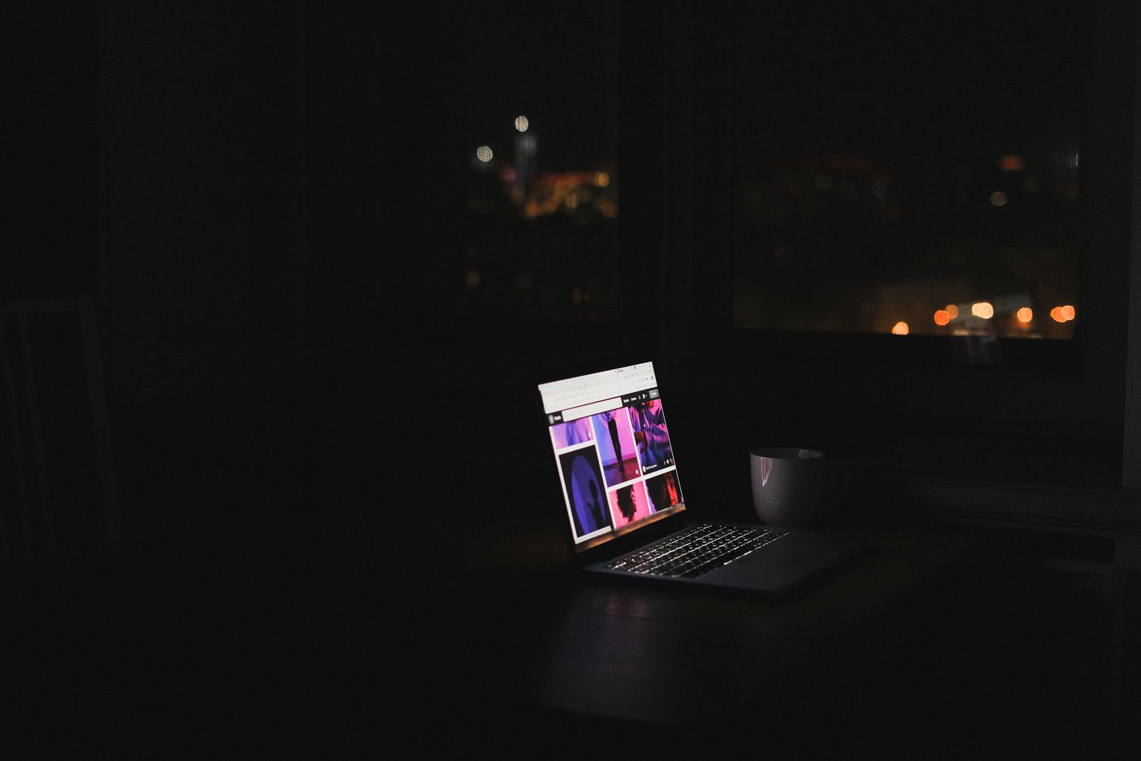 A laptop glowing on a desk in a dark room at night, city lights through the window