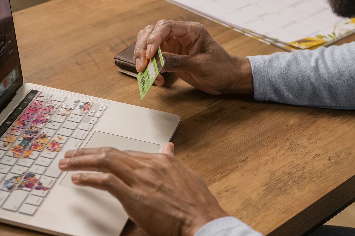 A person at a laptop on a wooden desk, holding a green payment card in one hand