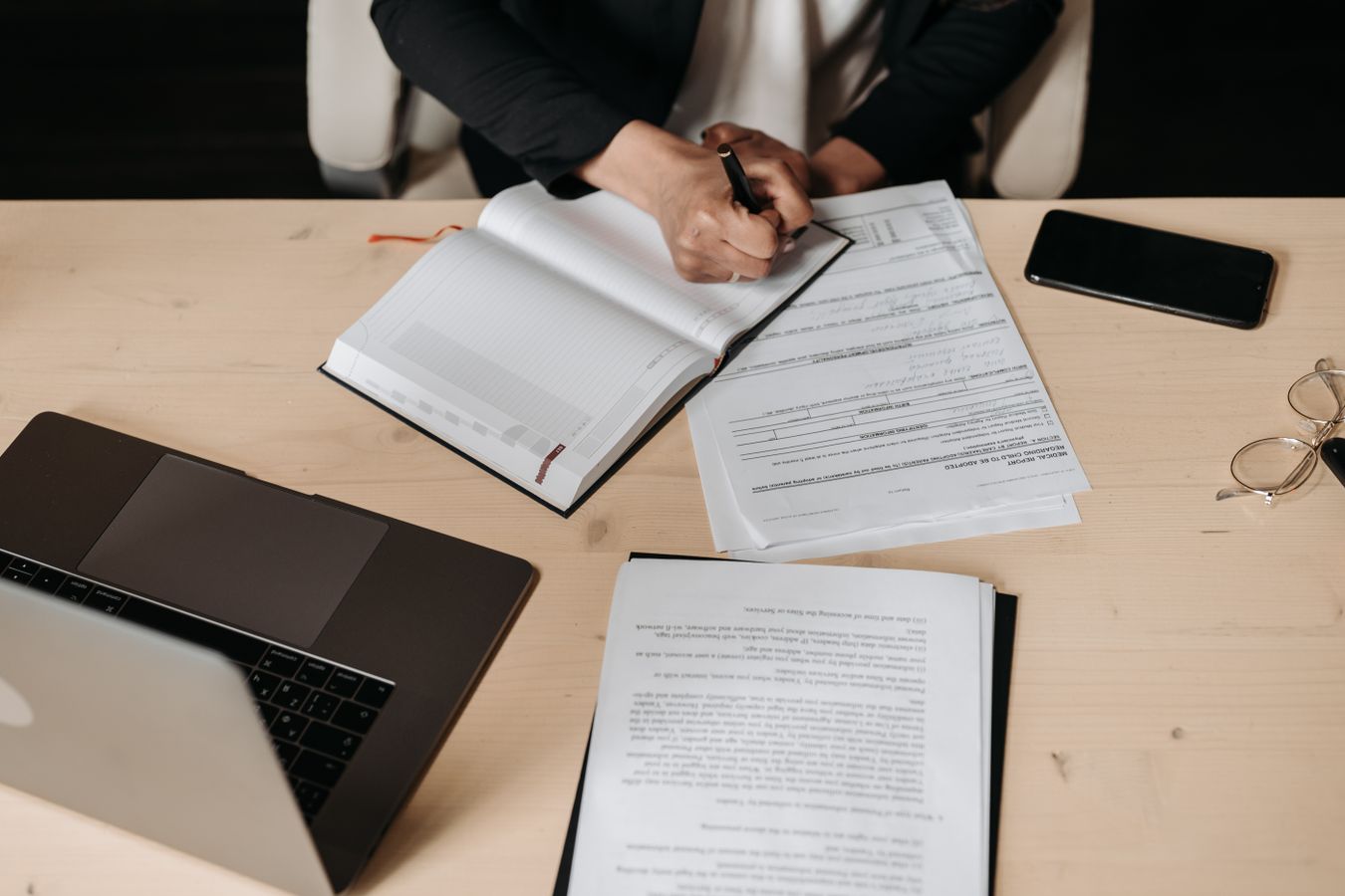 A person in a dark suit fills in printed forms at a wooden desk beside a laptop and a stack of papers