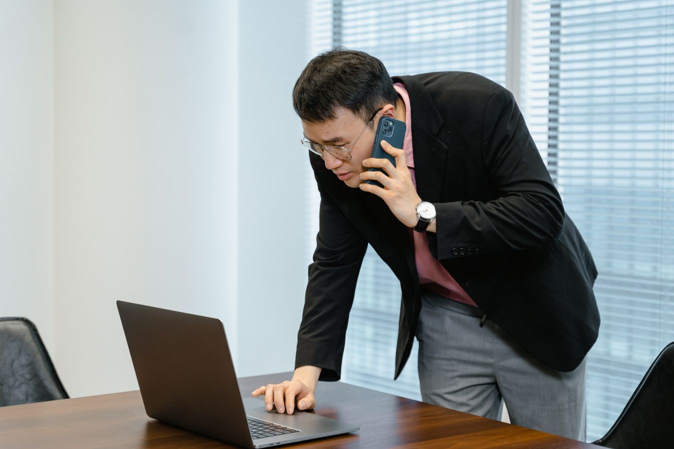 A man in glasses and a dark blazer leans over a conference table, phone pressed to his ear, frowning at an open laptop