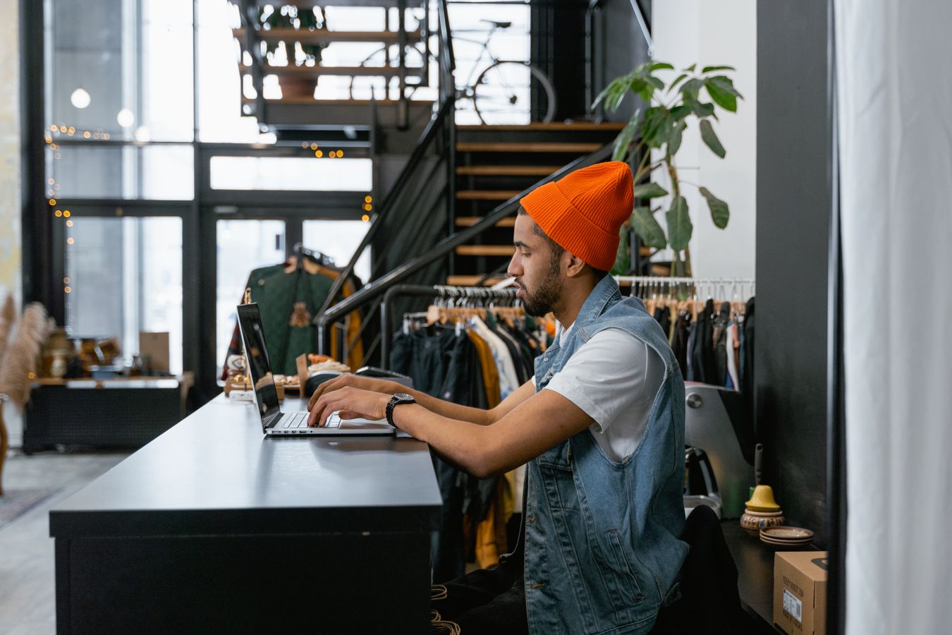 A shop owner in an orange beanie types on a laptop at the counter of a small clothing store