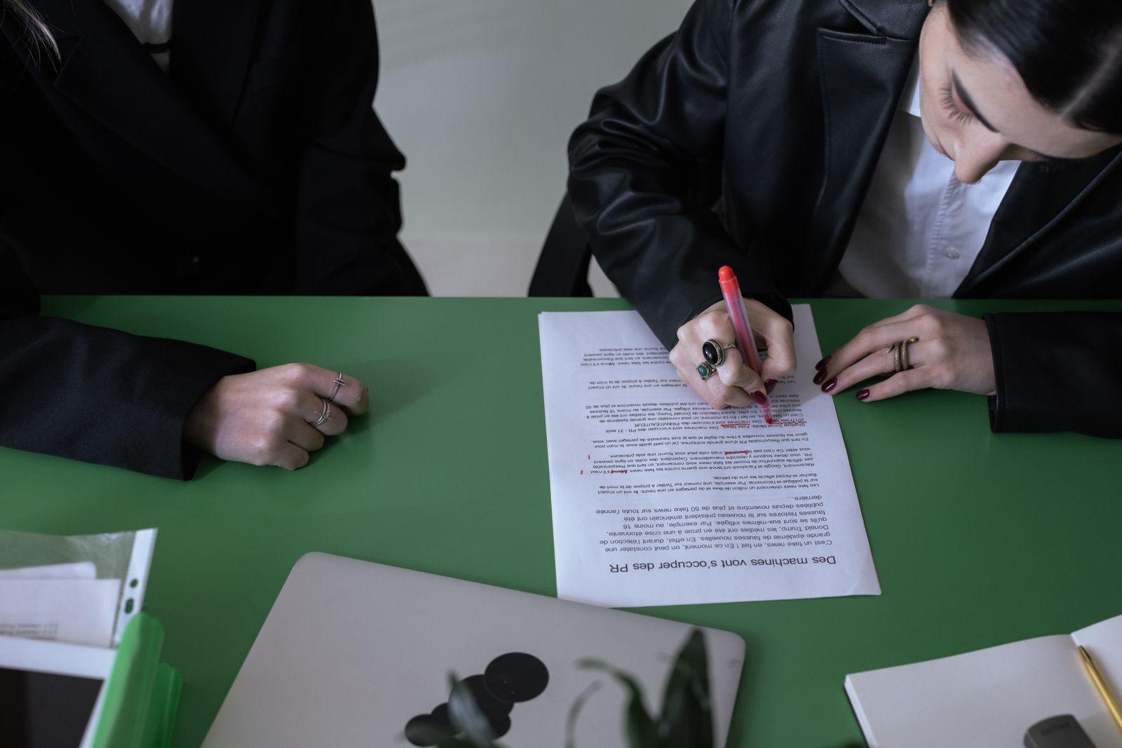 A person marking a printed page with a red pen at a green desk, a laptop beside it