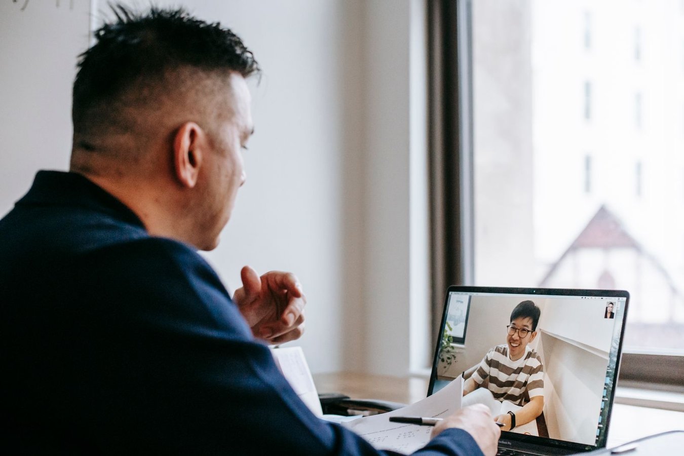 A man at a desk by a window on a video call with a smiling developer in a striped T-shirt