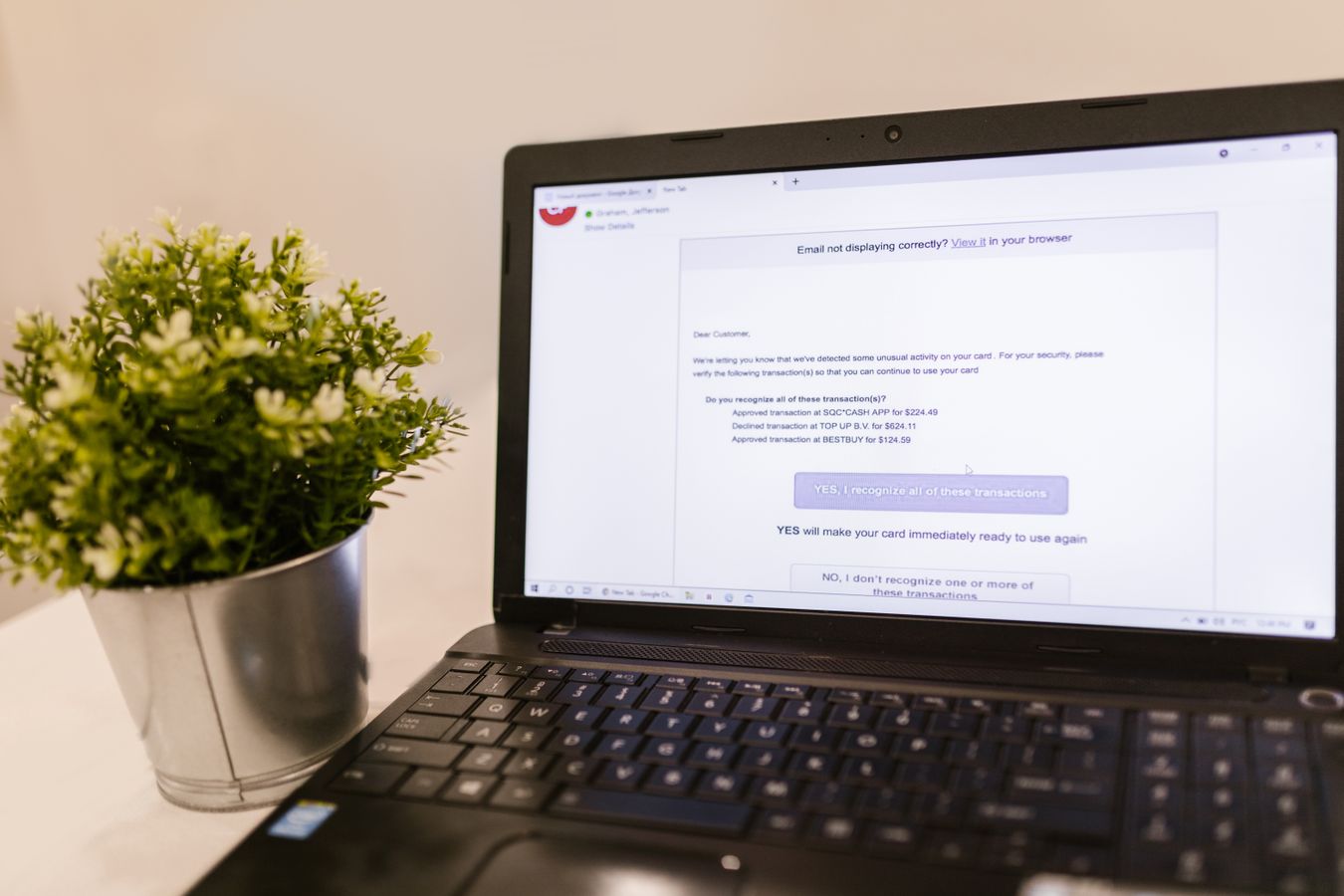 A laptop on a white desk showing an email that asks the reader to confirm card transactions, a small potted plant beside it