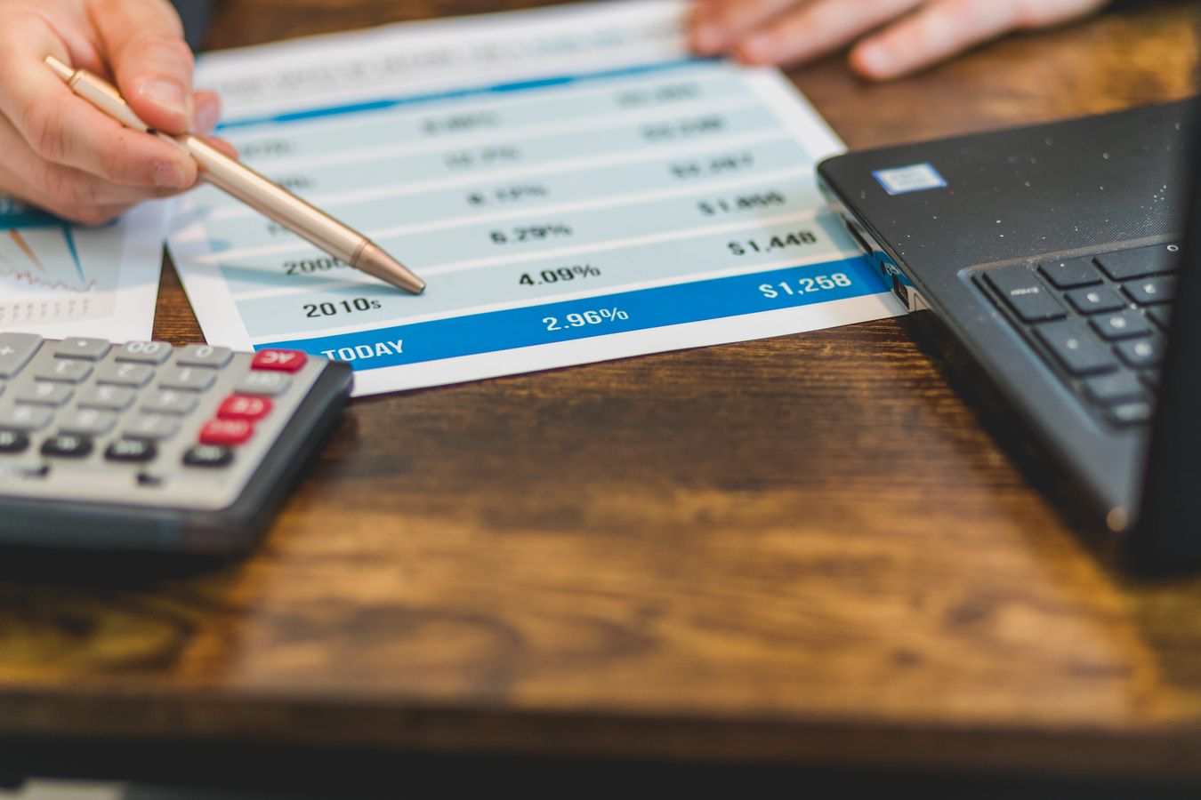 A pen pointing at a printed table of interest rates, a calculator and a laptop beside it on a wooden desk