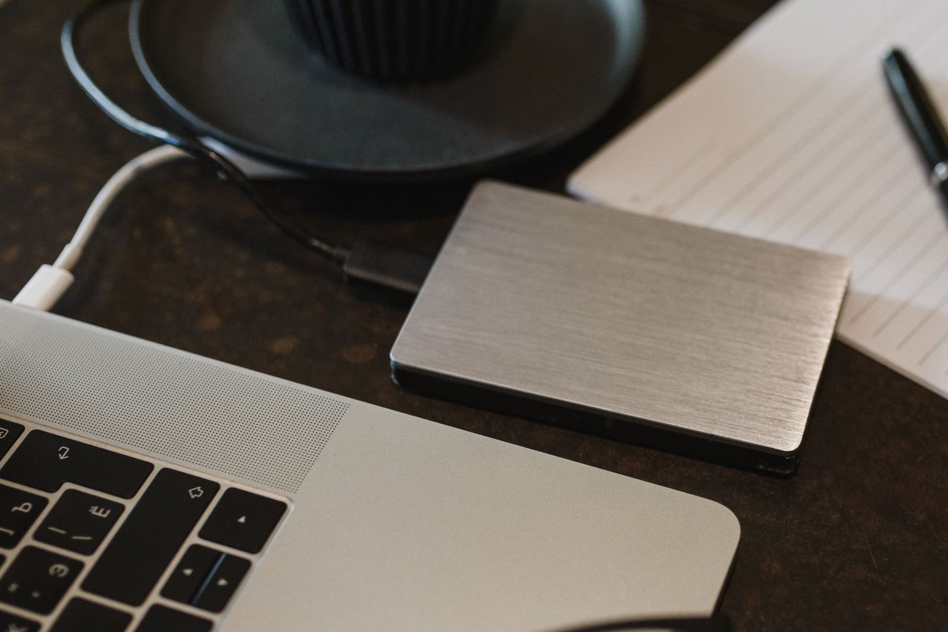 A brushed-metal portable drive on a dark desk beside an open laptop and a notepad