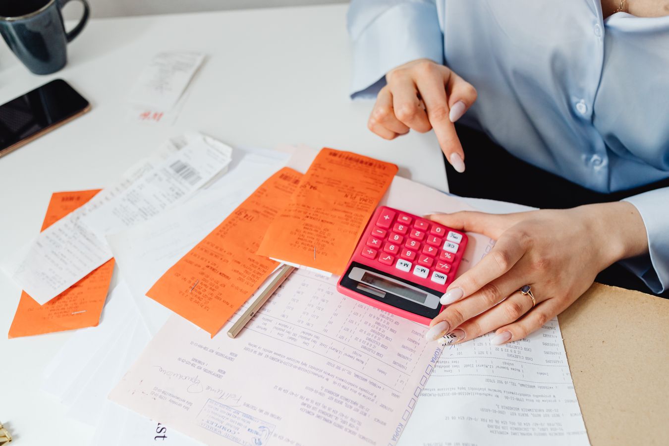 Hands on a pink calculator over orange receipts and printed invoices on a desk