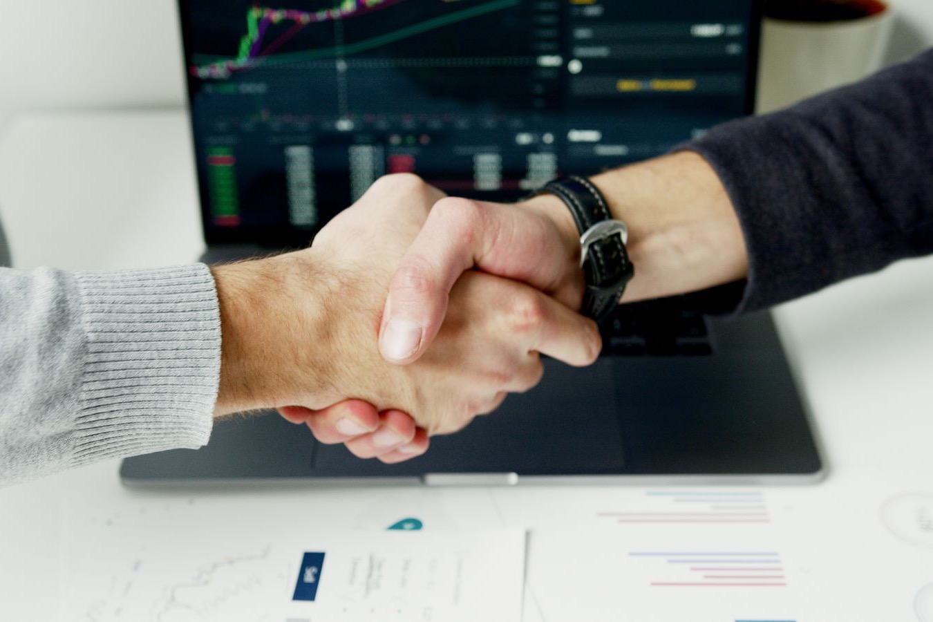 Two people shaking hands over a laptop showing financial charts, printed reports on the desk