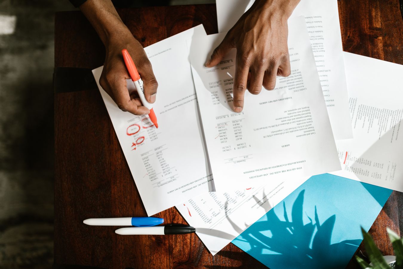 Two hands go through printed financial statements on a wooden desk, one circling figures with a red marker