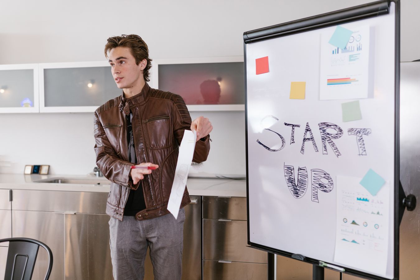 A young founder in a brown leather jacket presenting next to a flip chart with START UP written on it and charts pinned beside